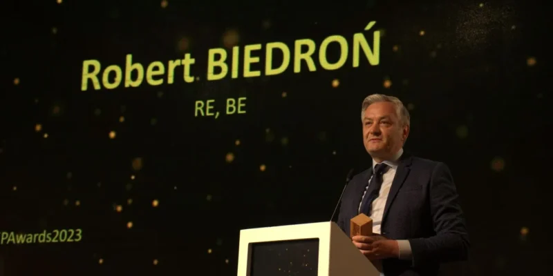 Mężczyzna w garniturze stoi przy podium na scenie podczas ceremonii, a w tle widoczny jest duży ekran z napisem i błyszczącymi punktami świetlnymi.