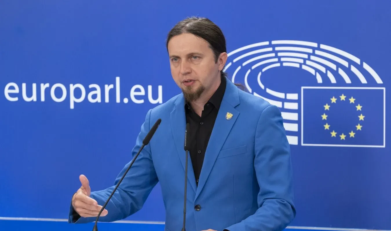 Mężczyzna z długimi włosami związanymi w kucyk i krótką brodą, ubrany w niebieski blazer, przemawia z mikrofonem przy podium na tle baneru z logo Unii Europejskiej.