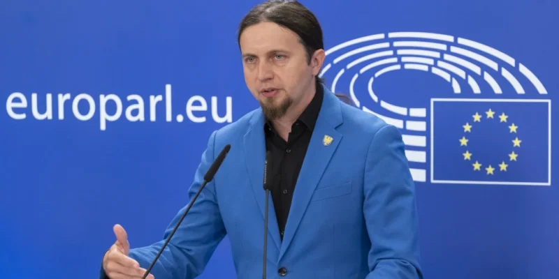 Mężczyzna z długimi włosami związanymi w kucyk i krótką brodą, ubrany w niebieski blazer, przemawia z mikrofonem przy podium na tle baneru z logo Unii Europejskiej.