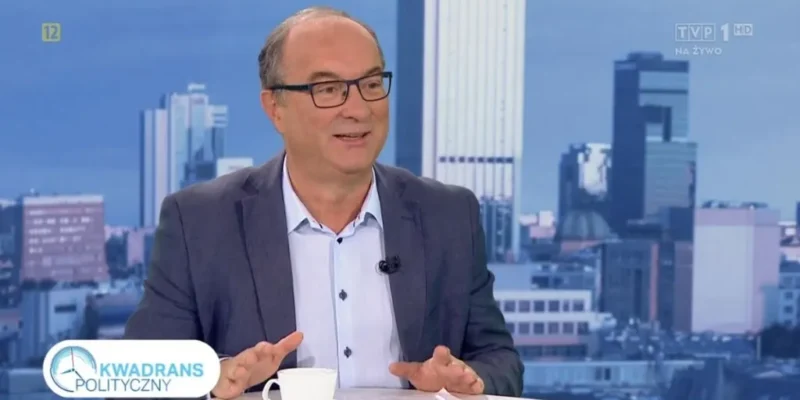 Mężczyzna w marynarce i okularach rozmawia w studiu telewizyjnym „Kwadrans Polityczny”, siedząc przy stole z filiżanką i dokumentami na tle panoramy miasta.