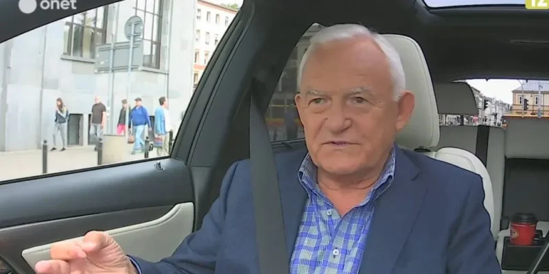 Starszy mężczyzna w marynarce, przypięty pasem, gestykuluje i udziela wywiadu w samochodzie na tle ulicy podczas rozmowy o reformie sądownictwa.