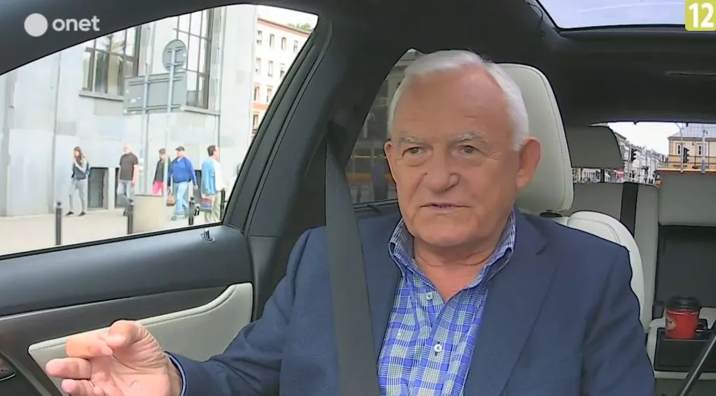 Starszy mężczyzna w marynarce, przypięty pasem, gestykuluje i udziela wywiadu w samochodzie na tle ulicy podczas rozmowy o reformie sądownictwa.