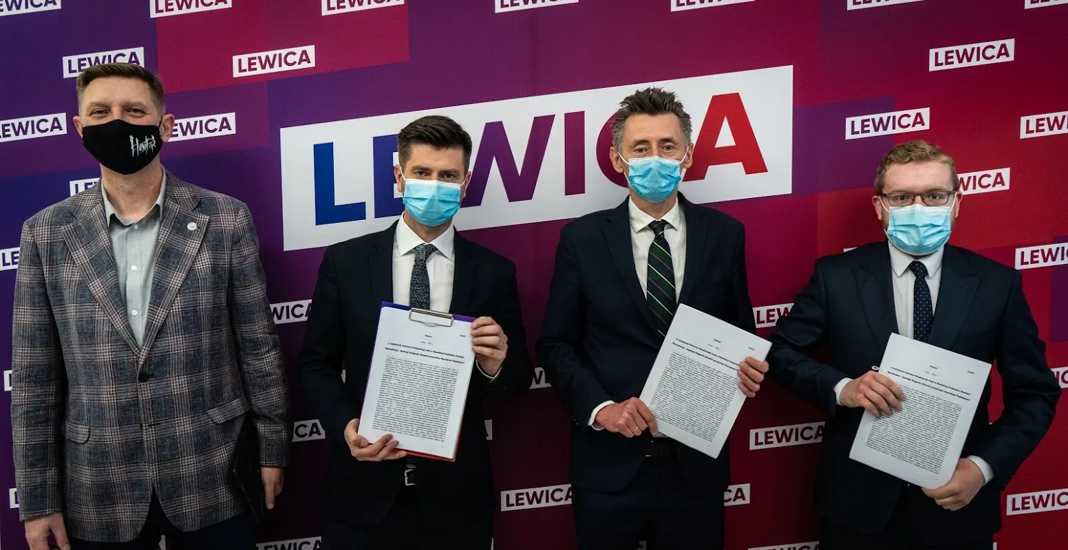 Na tle fioletowo-czerwonego tła z logo Lewica stoją czterech mężczyzn w garniturach i maseczkach, trzymających podpisane dokumenty.