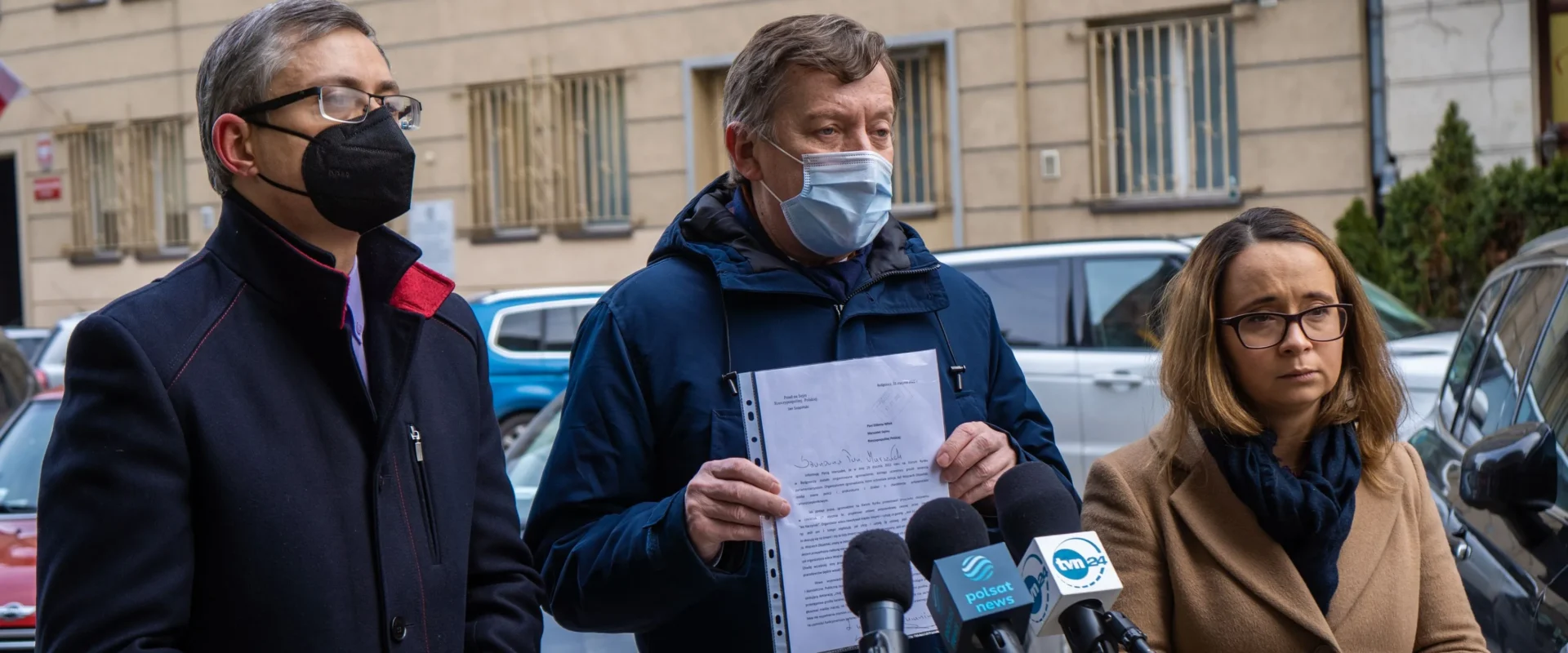 Na zdjęciu trzech mężczyzn i kobiet stoi przed budynkiem, trzymając dokumenty oraz rozmawiając z mikrofonami w tle.