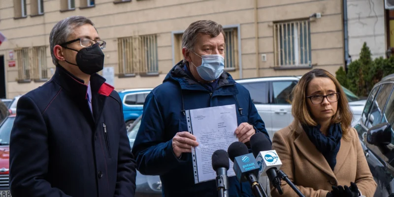 Na zdjęciu trzech mężczyzn i kobiet stoi przed budynkiem, trzymając dokumenty oraz rozmawiając z mikrofonami w tle.