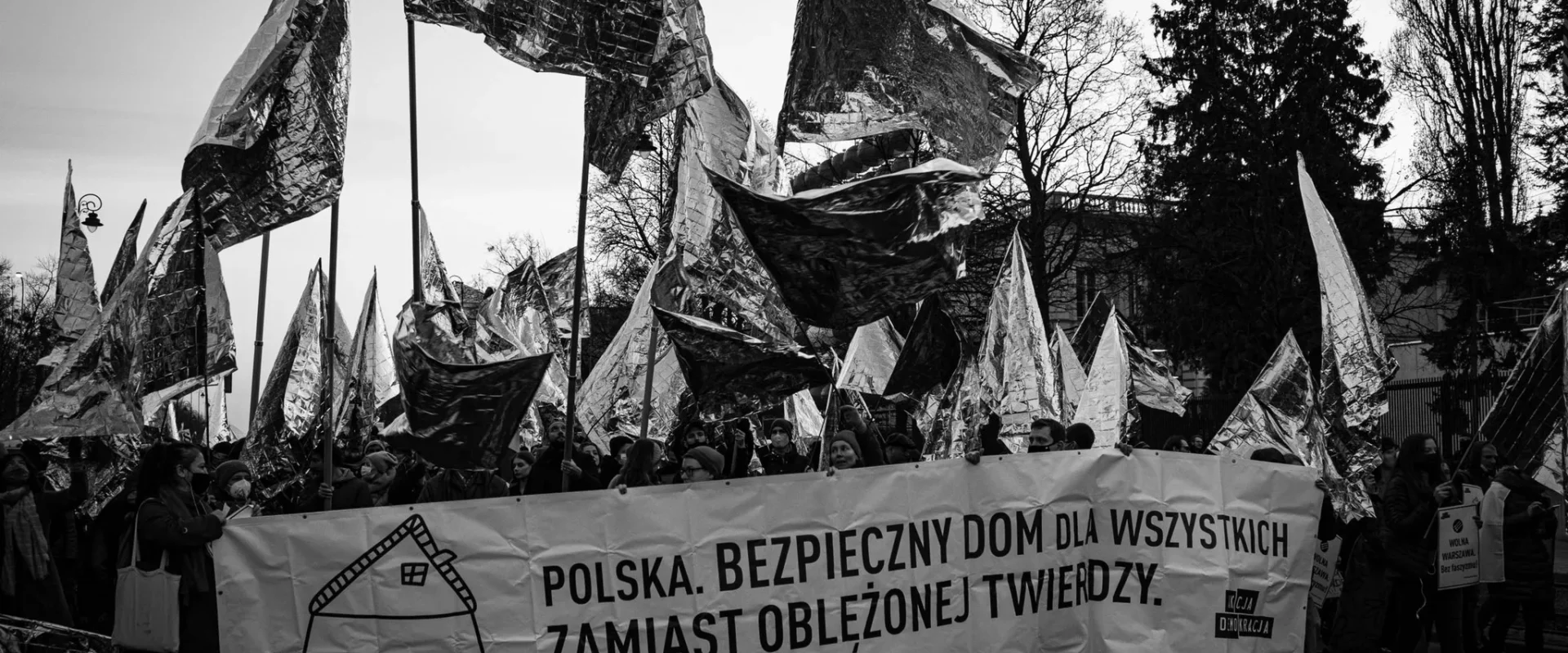 Na zdjęciu widoczny jest protest, na którym uczestnicy niosą transparent o treści "Polska. Bezpieczny dom dla wszystkich zamiast oblężonej twierdzy." oraz flagi w metalicznym kolorze.