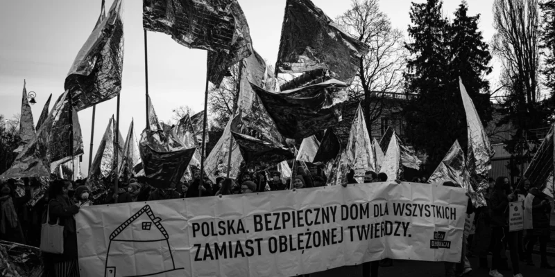 Na zdjęciu widoczny jest protest, na którym uczestnicy niosą transparent o treści "Polska. Bezpieczny dom dla wszystkich zamiast oblężonej twierdzy." oraz flagi w metalicznym kolorze.