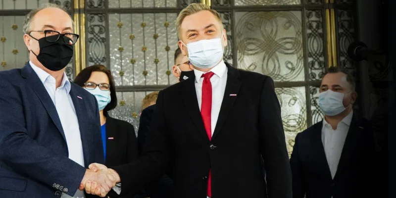 Dwoje mężczyzn w garniturach i maseczkach ściska sobie dłonie na tle ozdobnych drzwi, w tle stoją inni ludzie również w maseczkach.