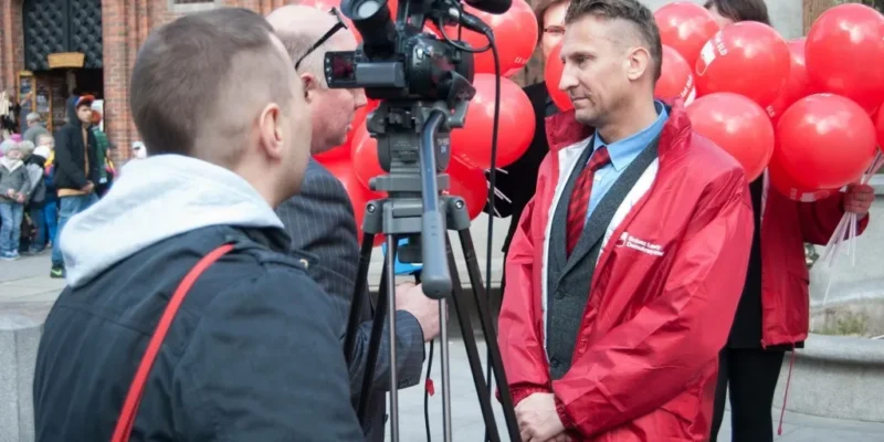 Mężczyzna w czerwonej kurtce stoi przed kamerą i mikrofonem, a w tle osoby trzymają czerwone balony na miejskim placu.