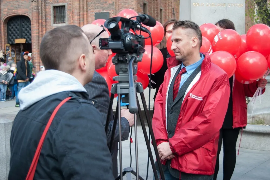 Mężczyzna w czerwonej kurtce stoi przed kamerą i mikrofonem, a w tle osoby trzymają czerwone balony na miejskim placu.
