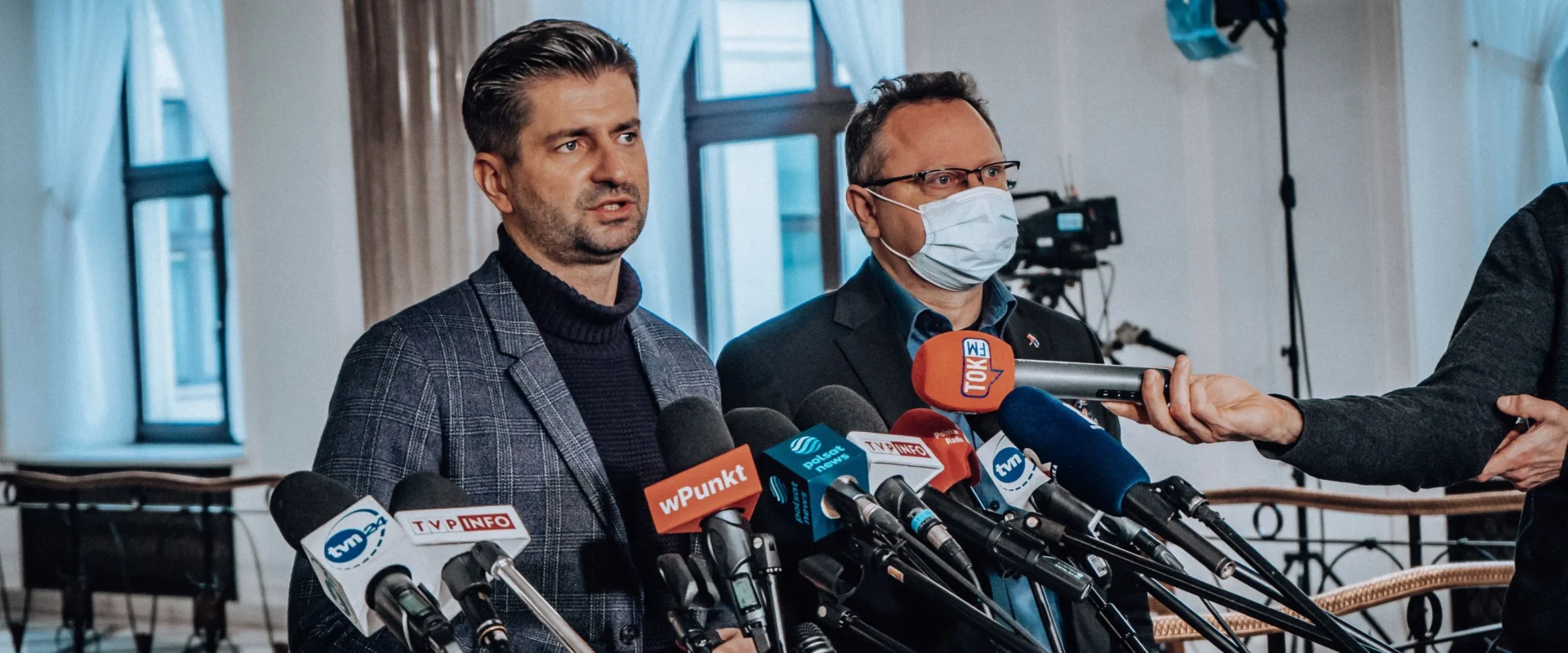 Na zdjęciu dwóch mężczyzn stoi przed mikrofonami w sali konferencyjnej, przygotowując się do udzielenia wywiadu.