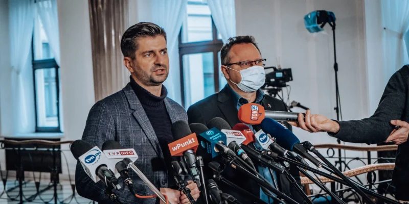 Na zdjęciu dwóch mężczyzn stoi przed mikrofonami w sali konferencyjnej, przygotowując się do udzielenia wywiadu.