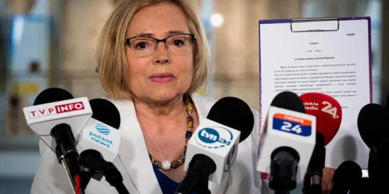 Kobieta w białej marynarce i okularach przemawia na konferencji prasowej, otoczona mikrofonami z logotypami stacji telewizyjnych.