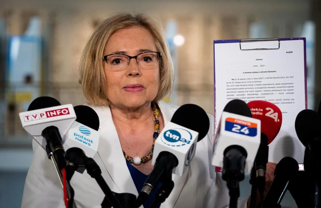 Kobieta w białej marynarce i okularach przemawia na konferencji prasowej, otoczona mikrofonami z logotypami stacji telewizyjnych.