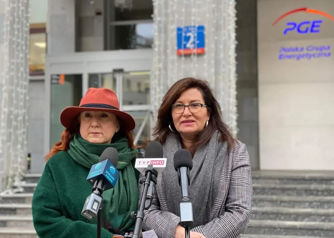 Na zdjęciu znajdują się dwie kobiety stojące przed siedzibą PGE, podczas konferencji prasowej, w jasnym otoczeniu ozdobionym świątecznymi światłami.