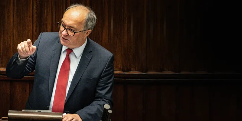 Mężczyzna w garniturze i czerwonym krawacie gestykuluje podczas przemówienia na tle drewnianych paneli.