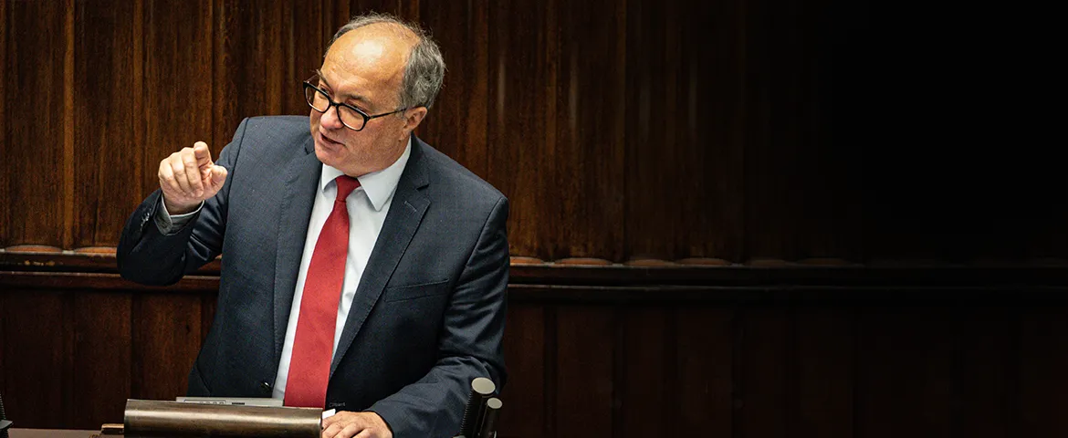 Mężczyzna w garniturze i czerwonym krawacie gestykuluje podczas przemówienia na tle drewnianych paneli.