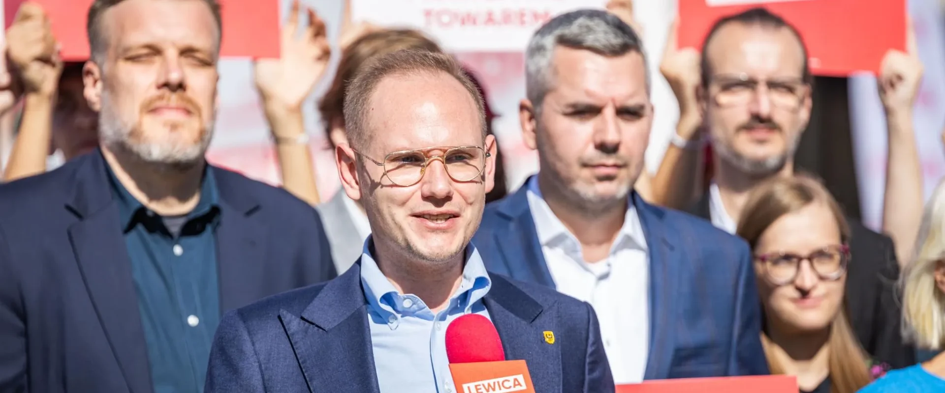 Mężczyzna w okularach w granatowym garniturze przemawia do mikrofonu z logiem Lewicy podczas wiecu, w tle grupa ludzi z czerwonymi plakatami Lewicy.