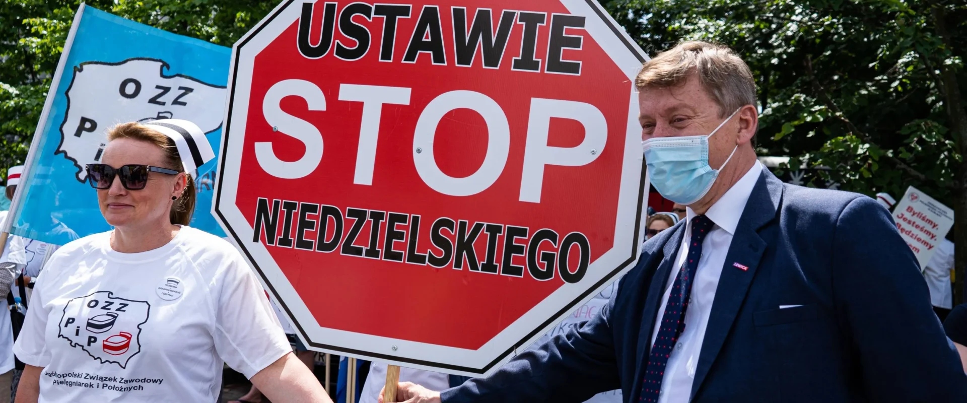 Dwóch protestujących – mężczyzna w garniturze i maseczce oraz kobieta w białej koszulce – trzymają duży czerwony znak STOP podczas demonstracji pielęgniarek i położnych.
