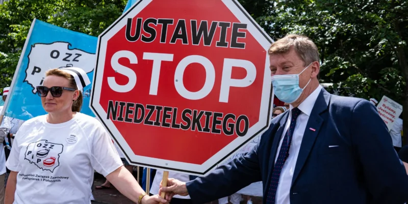 Dwóch protestujących – mężczyzna w garniturze i maseczce oraz kobieta w białej koszulce – trzymają duży czerwony znak STOP podczas demonstracji pielęgniarek i położnych.