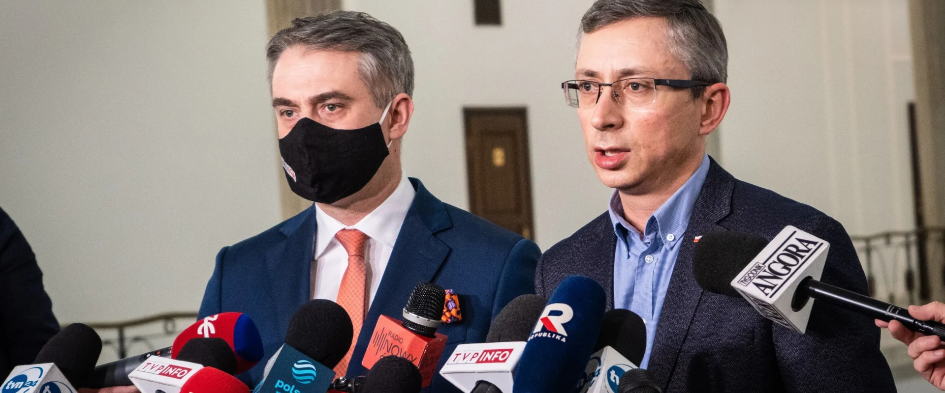 Na zdjęciu dwóch mężczyzn w garniturach, jeden z nich w maseczce, udziela wywiadu przed dziennikarzami trzymającymi mikrofony.