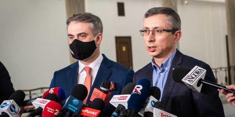 Na zdjęciu dwóch mężczyzn w garniturach, jeden z nich w maseczce, udziela wywiadu przed dziennikarzami trzymającymi mikrofony.