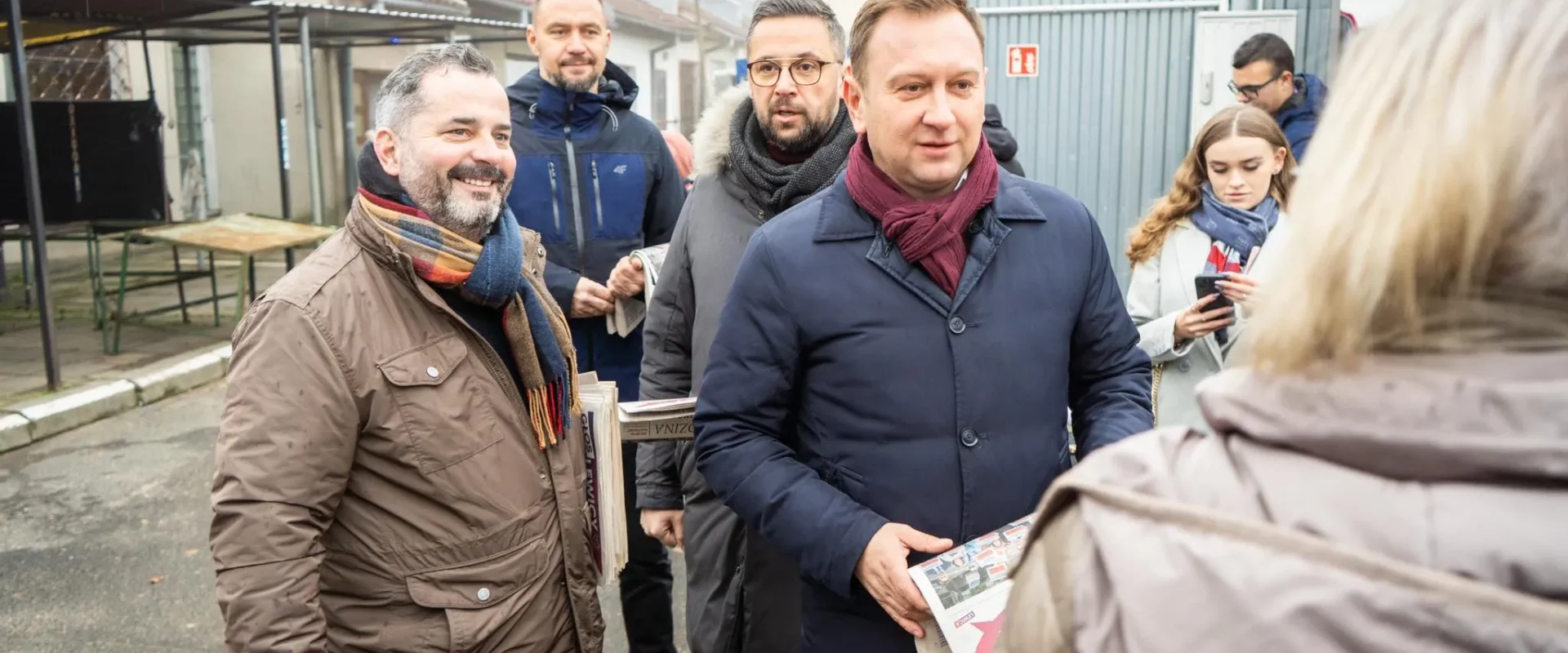 Grupa dorosłych w zimowych kurtkach stoi na ulicy podczas akcji „Bezpieczna Rodzina” na Dolnym Śląsku, rozdając gazety i ulotki.