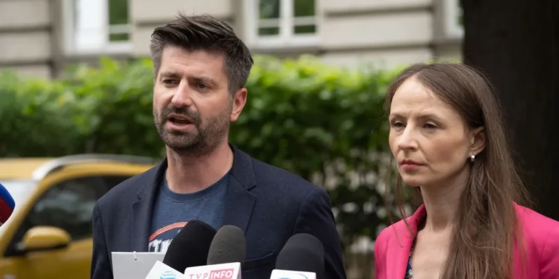 Mężczyzna w garniturze i kobieta w różowej marynarce stoją przed mikrofonami, wygłaszając oświadczenie w otoczeniu zieleni.