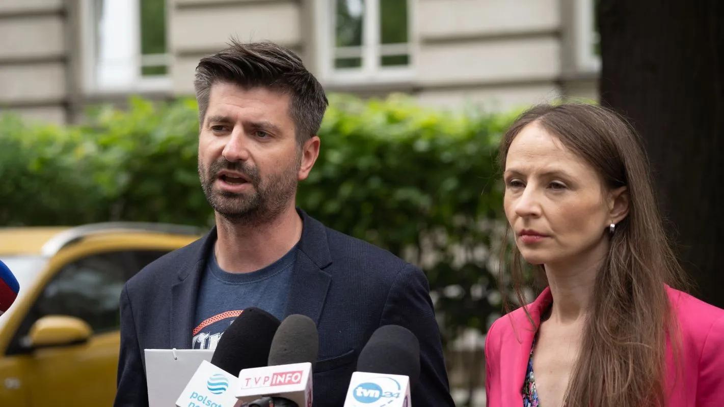 Mężczyzna w garniturze i kobieta w różowej marynarce stoją przed mikrofonami, wygłaszając oświadczenie w otoczeniu zieleni.