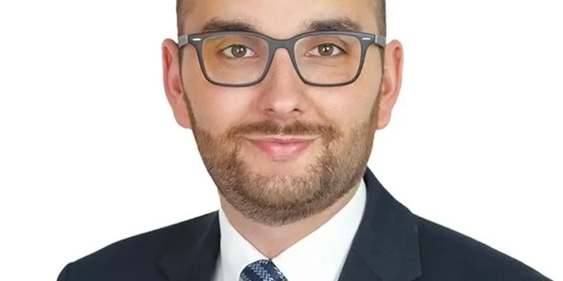 Mężczyzna w ciemnym garniturze i okularach, uśmiechnięty, na białym tle.