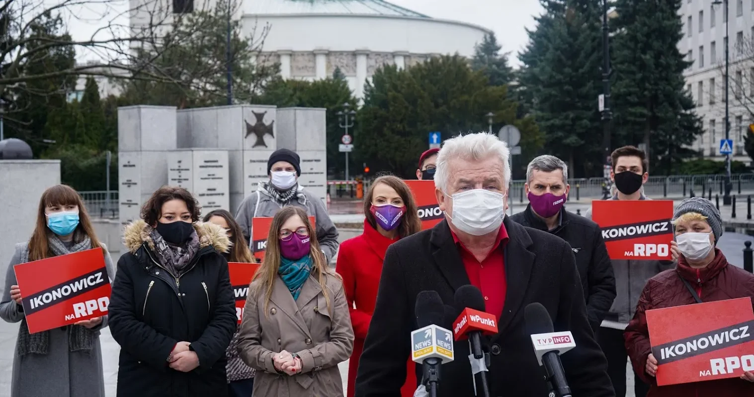Konferencja prasowa na zewnątrz: starszy mężczyzna w czerwonej koszuli i czarnym płaszczu przemawia do mikrofonów, stojąc przed grupą osób w maseczkach trzymających czerwone plakaty z napisem IKONOWICZ NA RPO.
