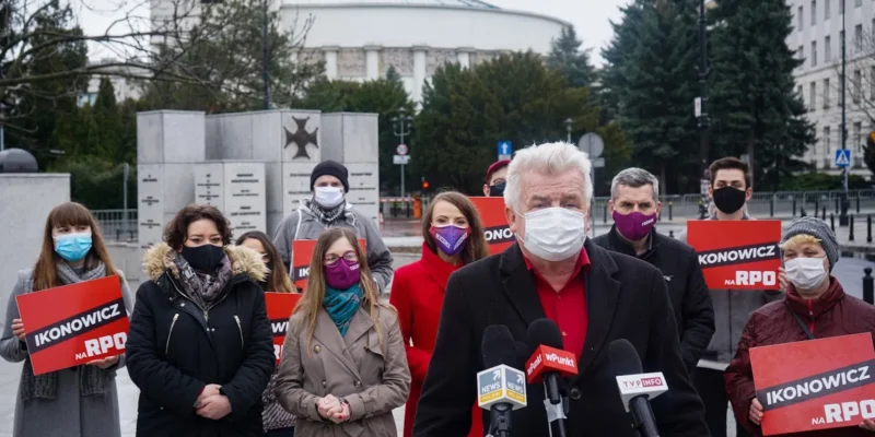 Konferencja prasowa na zewnątrz: starszy mężczyzna w czerwonej koszuli i czarnym płaszczu przemawia do mikrofonów, stojąc przed grupą osób w maseczkach trzymających czerwone plakaty z napisem IKONOWICZ NA RPO.