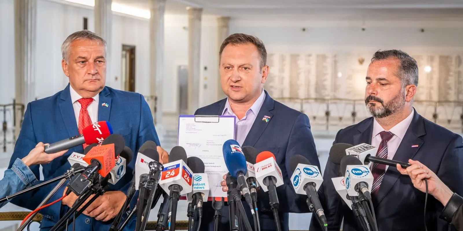 Trzech polityków podczas konferencji prasowej, jeden z nich trzyma dokument, a w tle widać mikrofony mediów.