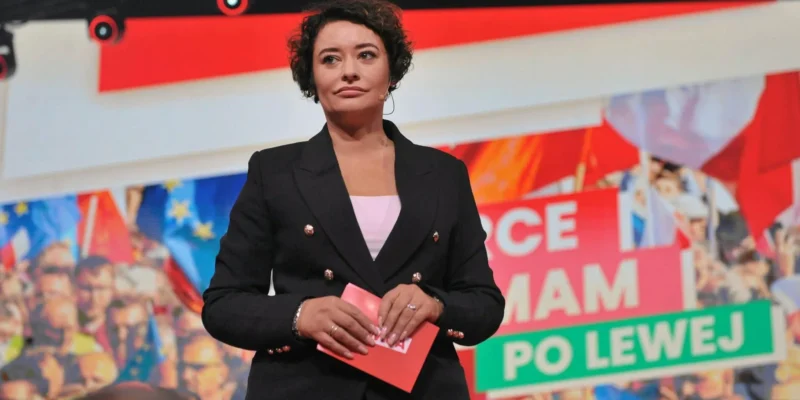 Kobieta w czarnym garniturze stoi na scenie z mikrofonem na uchu, trzyma czerwony notes, a w tle widoczne są kolorowe banery i tłum.