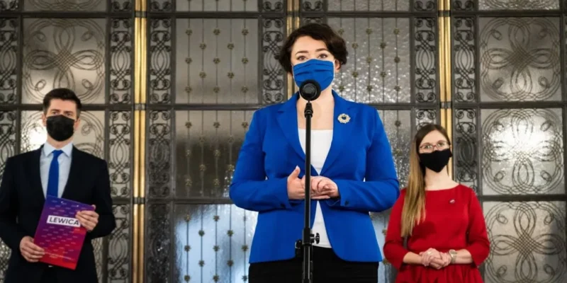 Trzy osoby stoją przed mikrofonem na tle ozdobnej ściany: pośrodku kobieta w niebieskim garniturze przemawia, z lewej mężczyzna w garniturze trzyma plik dokumentów, z prawej kobieta w czerwonej sukience obserwuje, wszyscy w maseczkach.