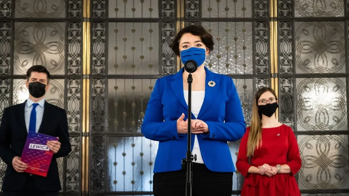 Trzy osoby stoją przed mikrofonem na tle ozdobnej ściany: pośrodku kobieta w niebieskim garniturze przemawia, z lewej mężczyzna w garniturze trzyma plik dokumentów, z prawej kobieta w czerwonej sukience obserwuje, wszyscy w maseczkach.