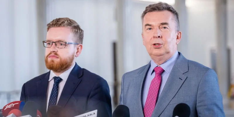 Dwóch mężczyzn w garniturach na konferencji prasowej stoi przed licznymi mikrofonami, z których jeden zabiera głos, drugi patrzy ku dziennikarzom.