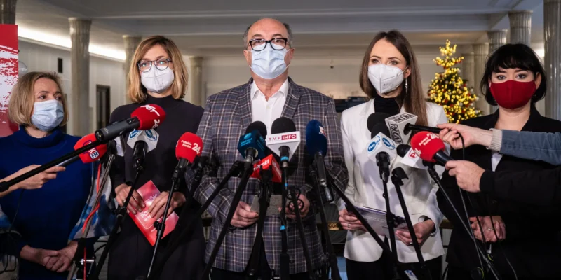 Grupa osób w maseczkach podczas konferencji prasowej na temat obowiązkowych szczepień na COVID-19, otoczona mikrofonami, z choinką w tle.