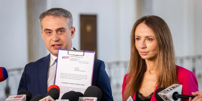 Dwoje polityków na konferencji prasowej stoi obok siebie przed licznymi mikrofonami, a mężczyzna trzyma dokument na clipboardie.