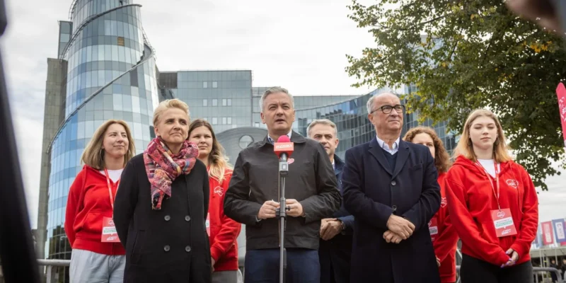 Grupa ludzi na zewnątrz przy nowoczesnym budynku, centralnie mężczyzna przemawia do mikrofonu, otoczony innymi uczestnikami w czerwonych bluzach.