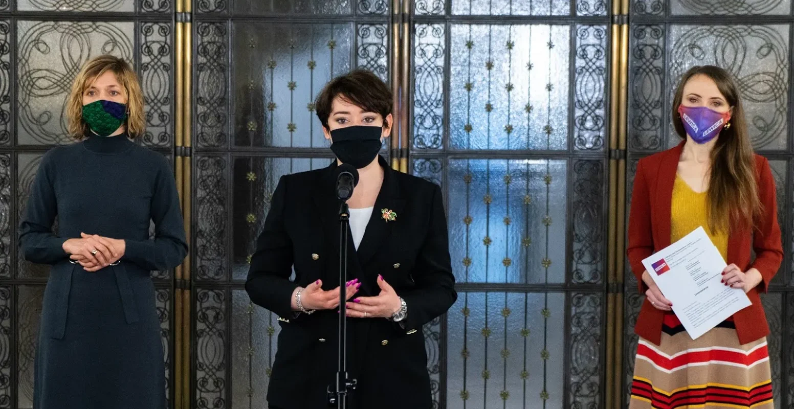 Trzy kobiety w maseczkach stoją przed ozdobnymi drzwiami; pośrodku kobieta wygłasza przemówienie przy mikrofonie, po prawej trzyma dokumenty.