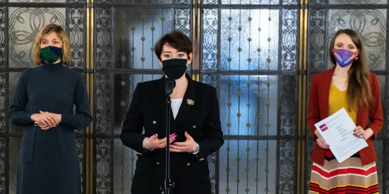 Trzy kobiety w maseczkach stoją przed ozdobnymi drzwiami; pośrodku kobieta wygłasza przemówienie przy mikrofonie, po prawej trzyma dokumenty.