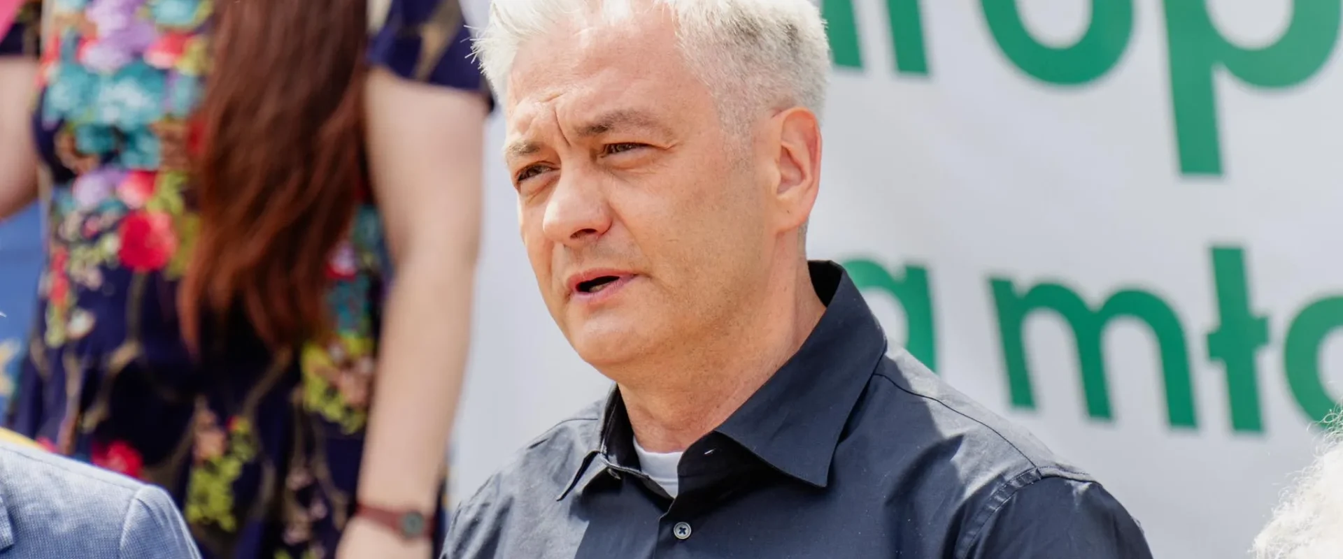 Mężczyzna o krótkich siwych włosach ubrany na czarno przemawia do mikrofonu podczas wydarzenia, w tle widoczny baner z zielonymi literami.