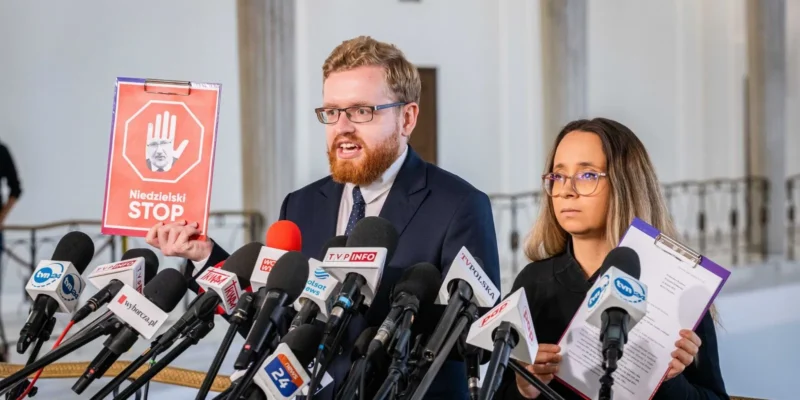 Mężczyzna z brodą i okularami w garniturze przemawia na konferencji prasowej, trzymając plakat z napisem „Niedzielski STOP”, obok stoi kobieta z teczką i notatkami, otoczony licznymi mikrofonami.