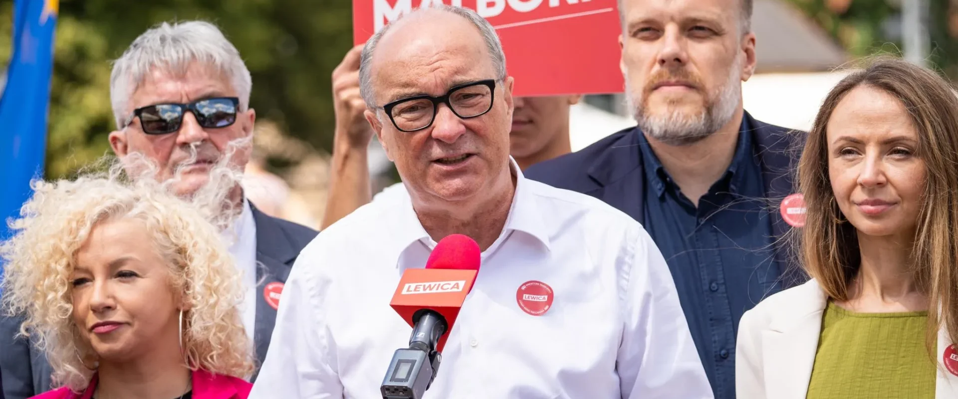 Grupa osób na wiecu politycznym Lewicy; mężczyzna w białej koszuli przemawia do mikrofonu z logo Lewicy, otoczony innymi uczestnikami i czerwonymi banerami w tle.