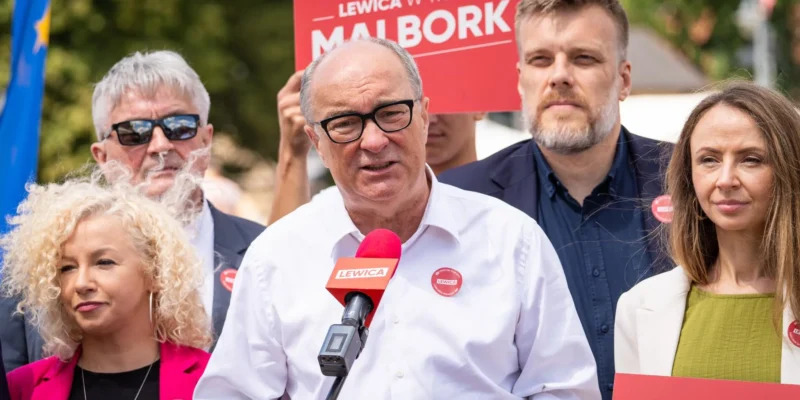 Grupa osób na wiecu politycznym Lewicy; mężczyzna w białej koszuli przemawia do mikrofonu z logo Lewicy, otoczony innymi uczestnikami i czerwonymi banerami w tle.