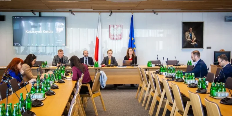 Zdjęcie przedstawia spotkanie w sali konferencyjnej, w której siedzi grupa osób przy długim stole, a na monitorze widać uczestnika zdalnego.