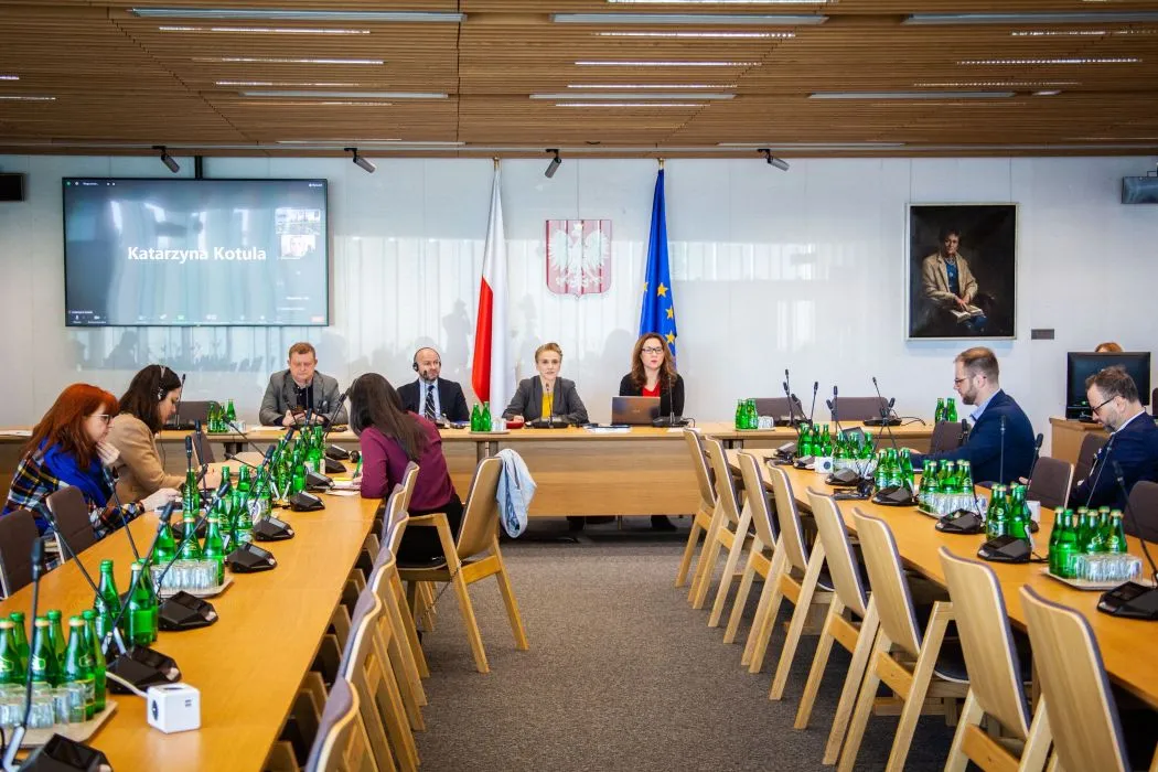 Zdjęcie przedstawia spotkanie w sali konferencyjnej, w której siedzi grupa osób przy długim stole, a na monitorze widać uczestnika zdalnego.