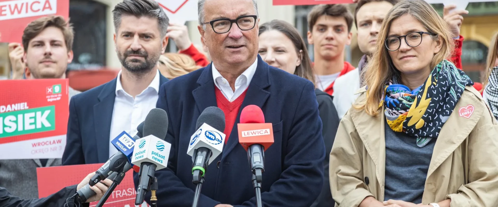 Polityk w okularach prowadzi konferencję prasową, stojąc przed zestawem mikrofonów, otoczony zwolennikami i plakatami Lewicy w tle.