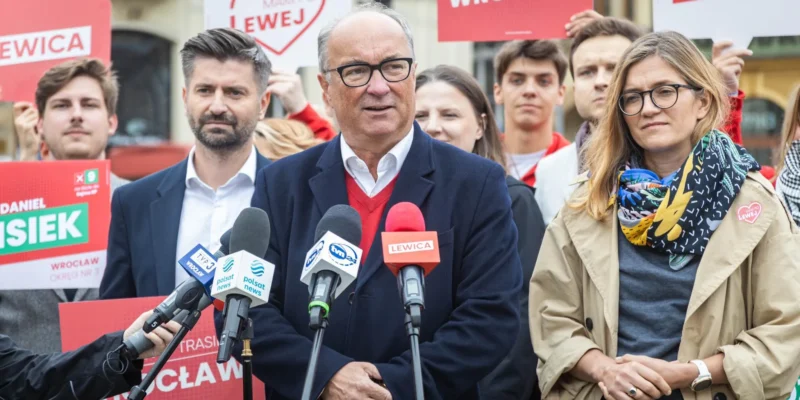 Polityk w okularach prowadzi konferencję prasową, stojąc przed zestawem mikrofonów, otoczony zwolennikami i plakatami Lewicy w tle.